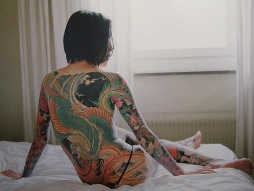 Woman sitting on a bed naked with her back covered in tattooes.