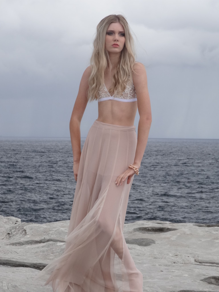 Photograph of Elise Faust standing by the ocean in a long beige see-through skirt and bikini top.