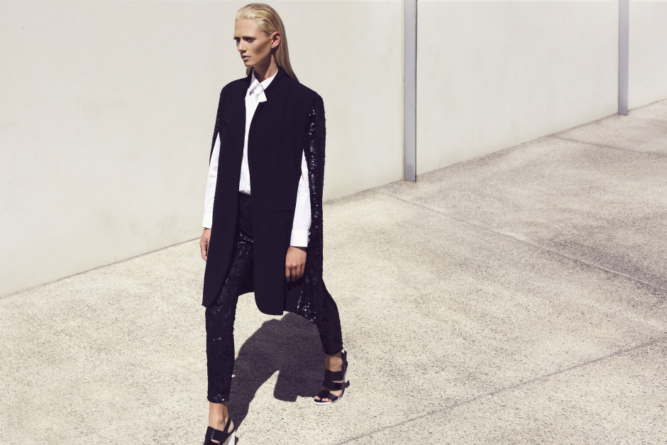 Model walking and being photographed in the full sunlight against a white wall wearing a black pant and black winter jacket with white shirt and heels