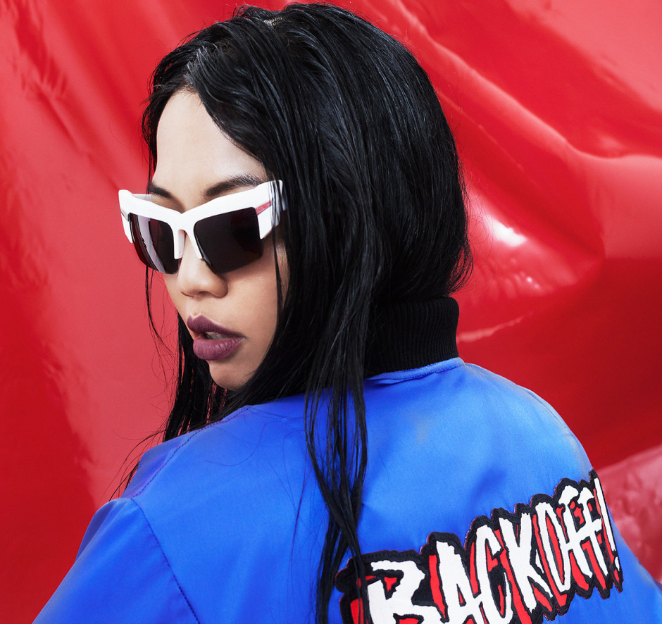 Autumn/Winter 2015. Asian model sitting in front a red vinyl sheet in a blue bomber jacket with writing across the back of the jacket. The model is being photographed in white mirrored sunglasses.