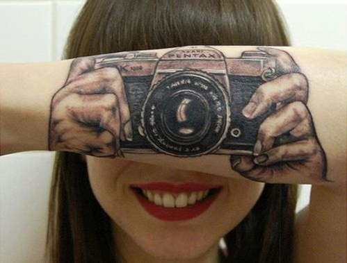 Girl smiling looking at the camera with her arm folded in front of her face with a tattoo on her arm of two hands holding a picture. So it looks like she is taking a picture of her arm.