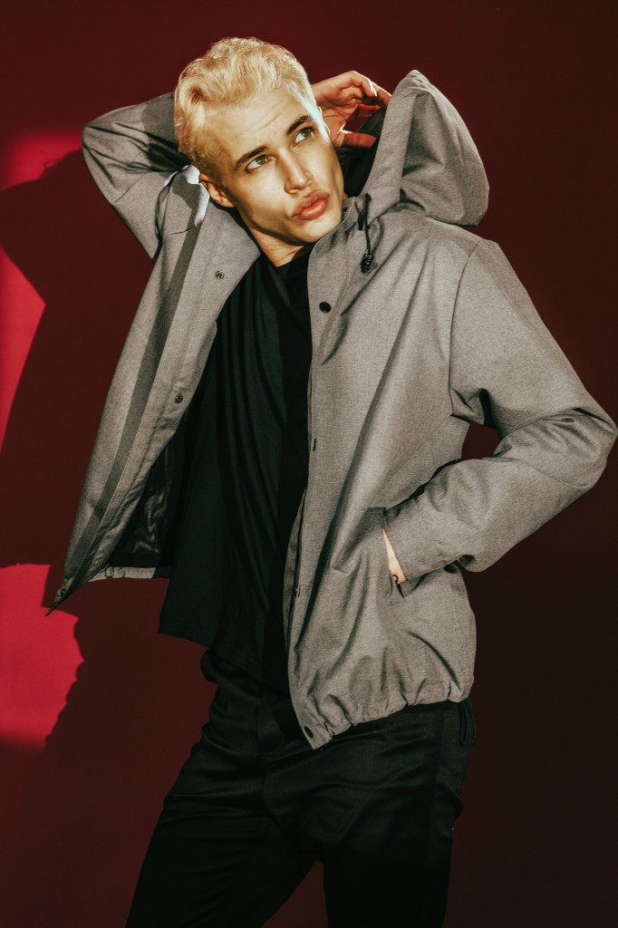 Model standing in front of a red wall dressed completely in black but wearing a donkey brown jacket with hoodie.