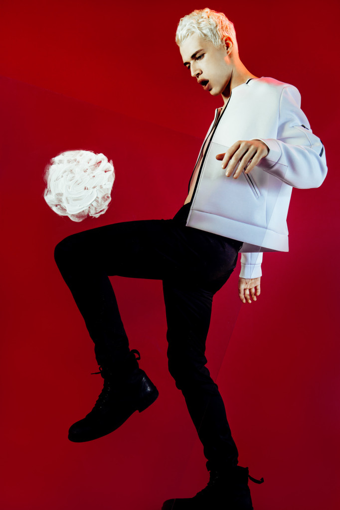 Model standing in front of a red wall kicking a virtual white ball above his knees.