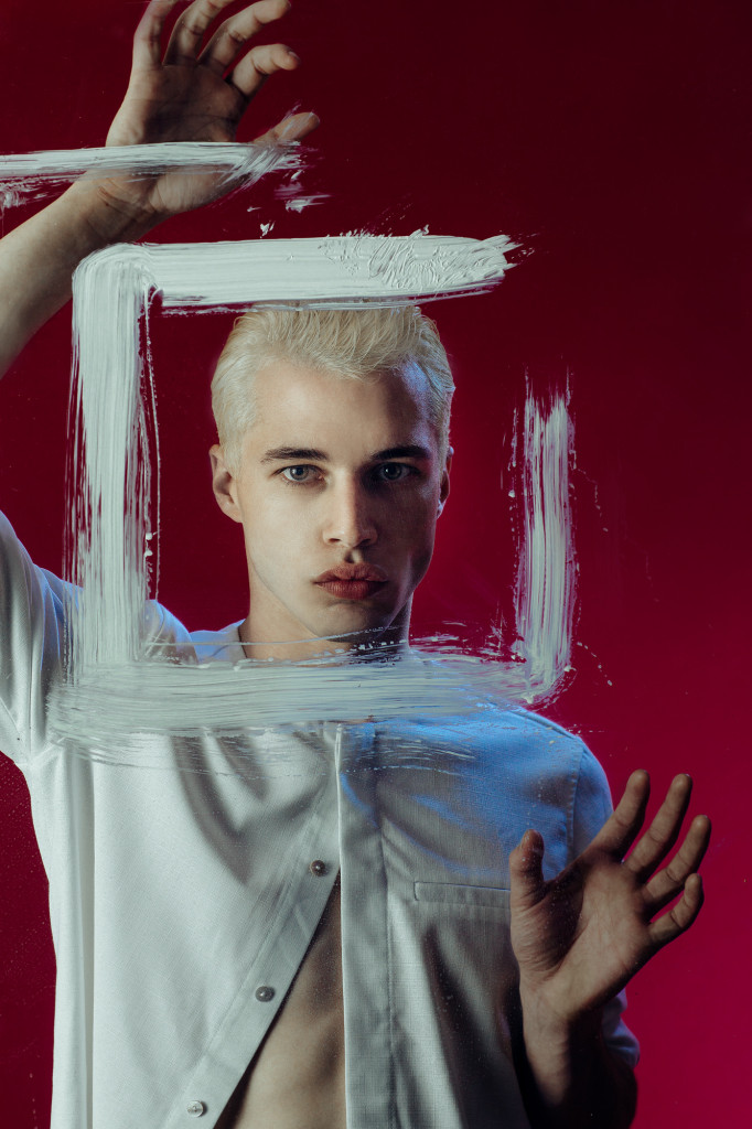 Model standing in front of a red wall dressed in a white t and facing the camera looking through clear perspex and a frame of white paint.