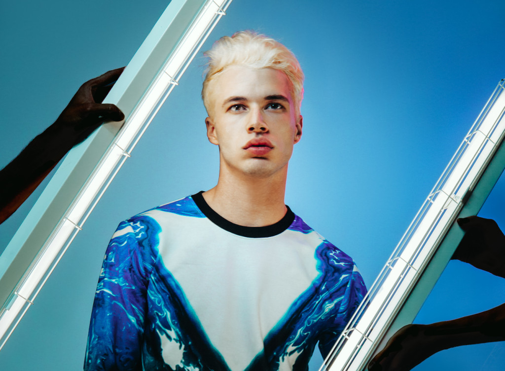 Male model standing in front of a blue wall in white t but with colourful blue tones jacket and two fluorescent tubes being held by hands and arms on either side of him.