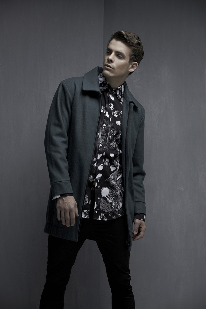 Model standing in front of a grey wall in a grey jacket, patterned shirt and black pants.