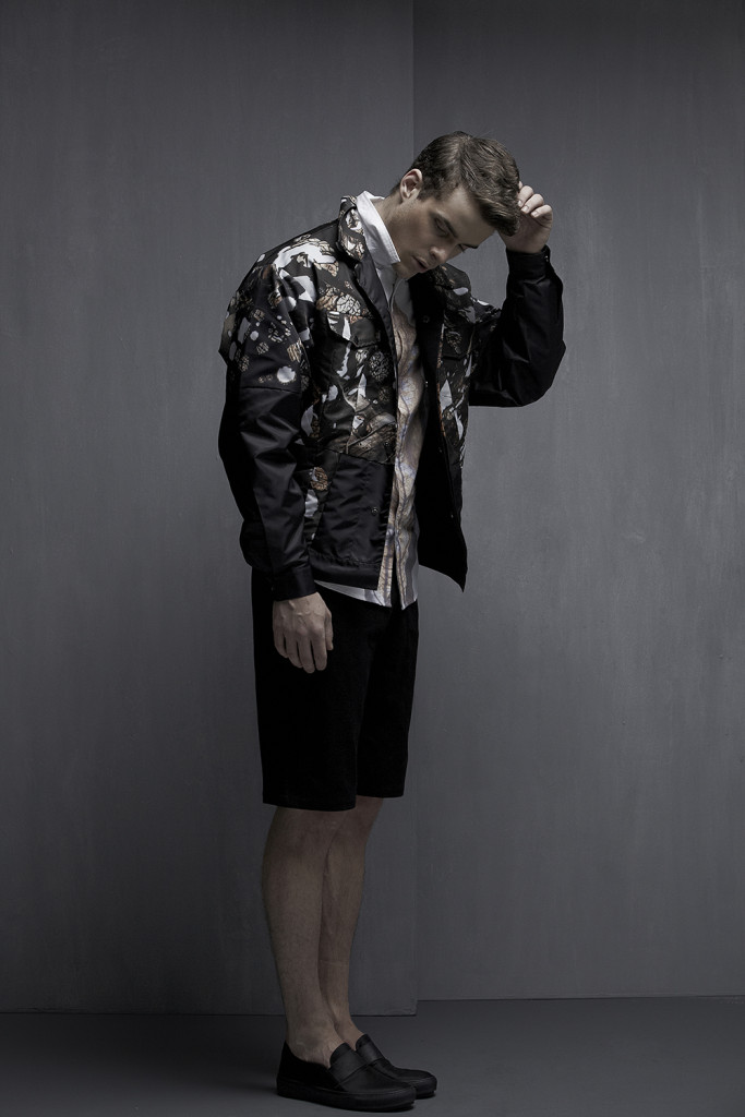 Model standing in front of a grey wall in a patterned jacket, black shorts and loafers.