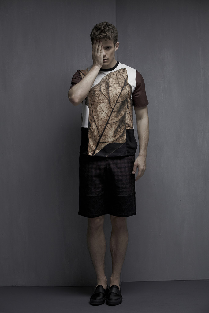 Male model standing in front of a grey wall in black shorts and a patterned T with short sleeves and a rust leaf motif on the front.