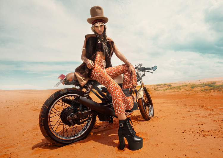 Girls sitting with high heeled black boots, bare legs, leather jacket, and hat sitting on a motor bike on a sandy beach.