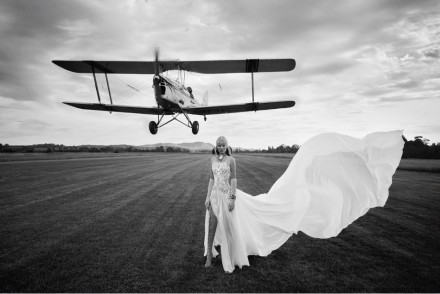 Model standing on an airfield in a beautiful white flowing dress, completely lace up open boots and vintage beaded head gear with an old fashioned aircraft taking off the background behind her. Very hollywood setting and incredibly creative the a real feel of movement to the picture.