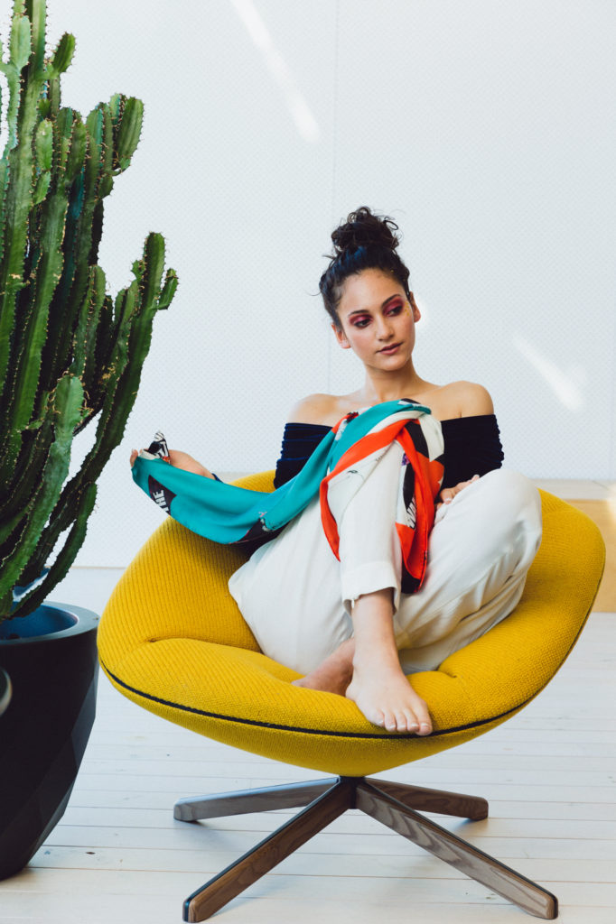 Model sitting for a photographic shoot for designer scarf label Valentina Avramides wearing a black open shoulder top, white pants on a bright yellow chair.