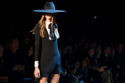 Model in a black dress and black hat walking the runway in front of an audience