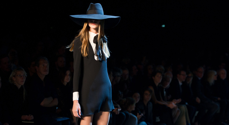 Model in a black dress and black hat walking the runway in front of an audience