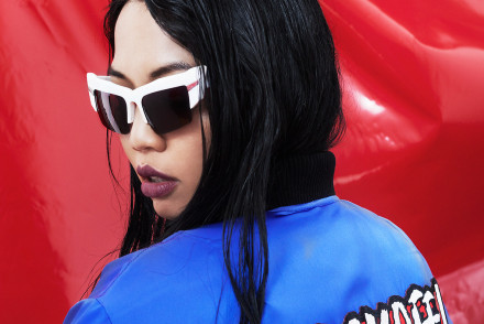 Autumn/Winter 2015. Asian model sitting in front a red vinyl sheet in a blue bomber jacket with writing across the back of the jacket. The model is being photographed in white mirrored sunglasses.